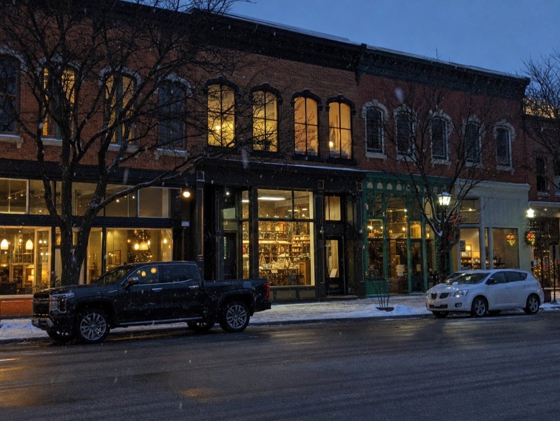 Independent businesses lit up in red-brick buildings on a small-town main street at dusk