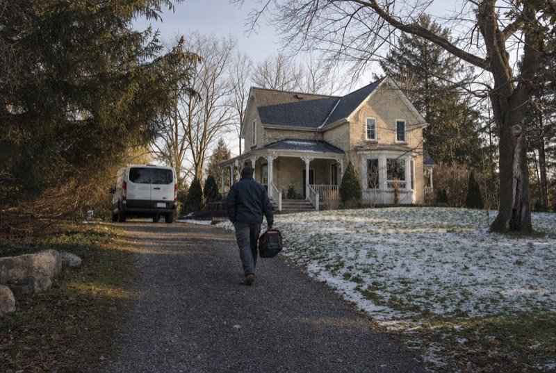 A technician carrying a toolbox up to a farmhouse-style home in early winter